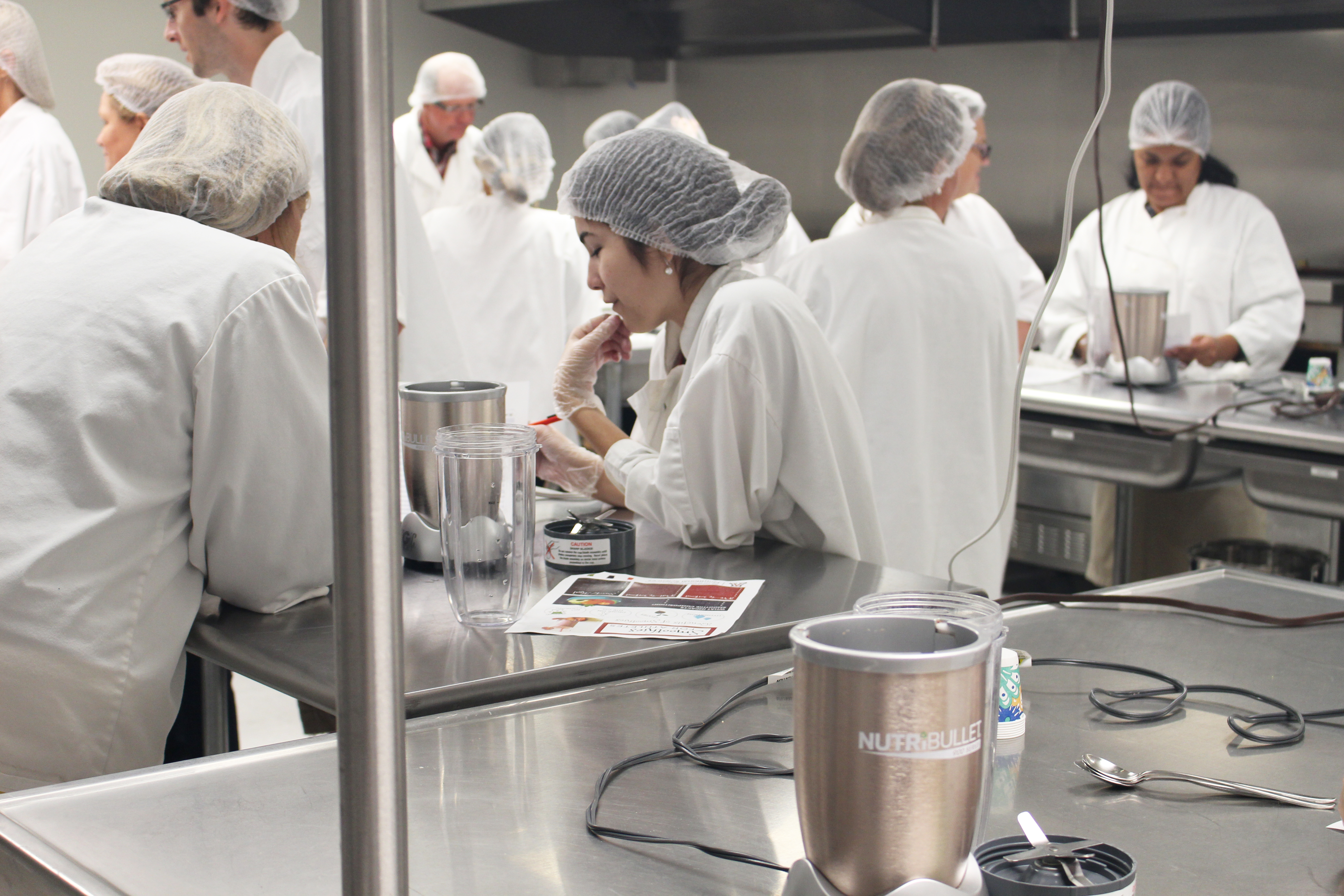 A room full of college students and faculty wearing white coats and hair nets work in an industrial kitchen.