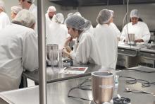 Several college students wearing white coats and hair nets work in an industrial kitchen.