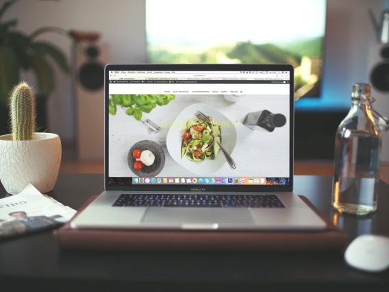 A silver laptop displaying a full-screen image of healthy food sits on a black desktop, facing the camera. To the left of the laptop is a potted cactus and a folded newspaper. To the right is a clear, glass water bottle.