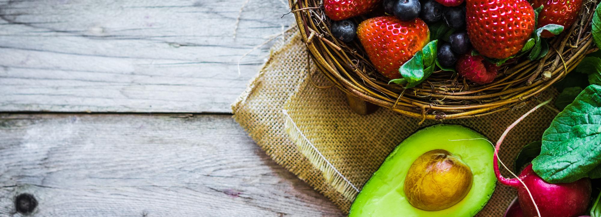 Close-up of a basket with berries, half an avocado, and a bunch of radishes on a wooden table.