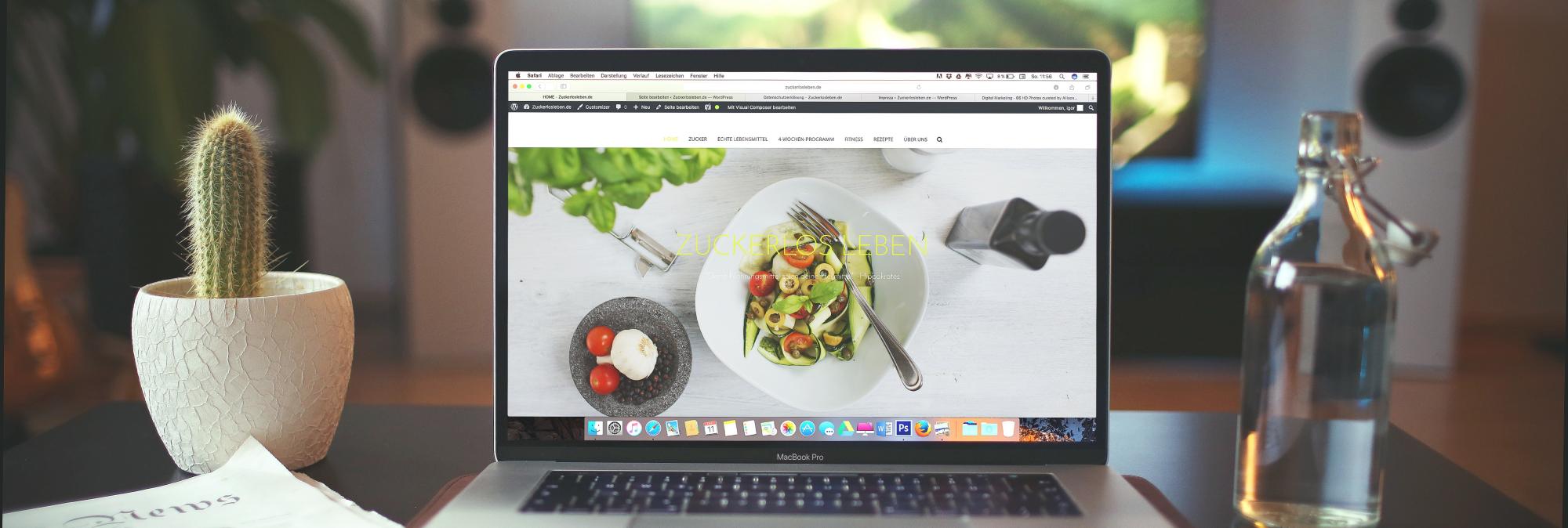 A silver laptop displaying a full-screen image of healthy food sits on a black desktop, facing the camera. To the left of the laptop is a potted cactus and a folded newspaper. To the right is a clear, glass water bottle.